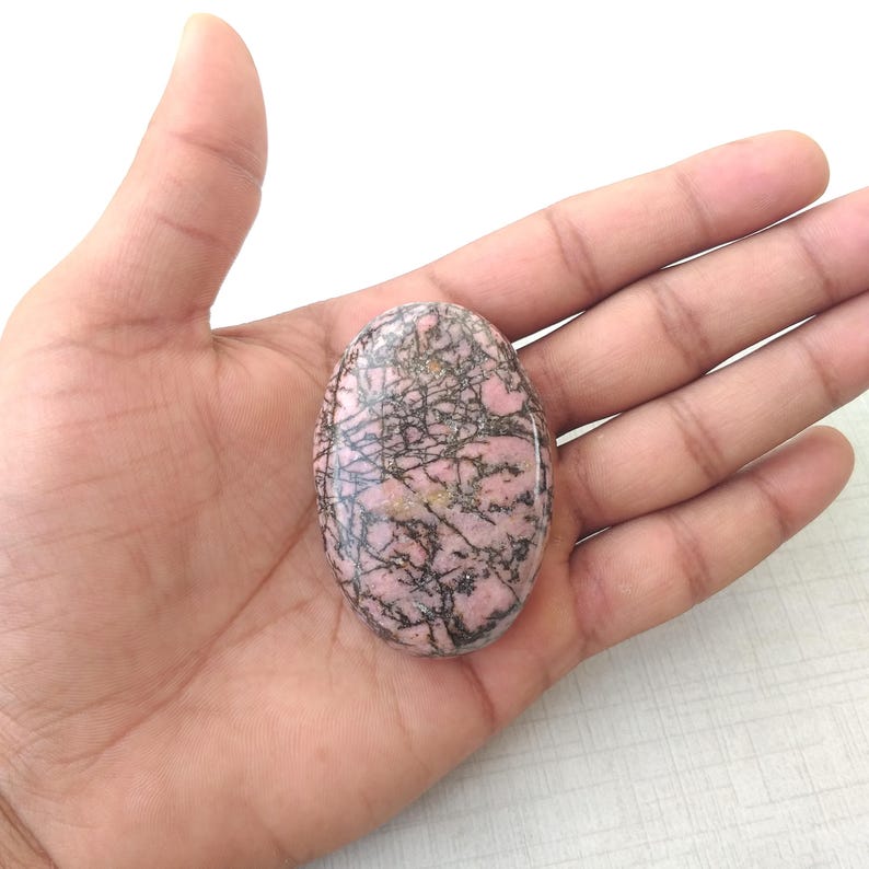May include: Oval-shaped rhodonite gemstone with a pink base and black veining. The polished stone is held in an open hand, showcasing its natural patterns and colors. This gemstone is a unique piece for jewelry or collection.