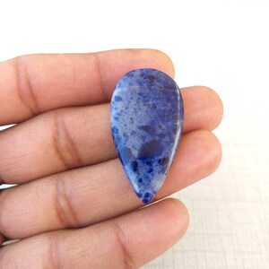 May include: A teardrop-shaped, polished gemstone with a deep blue hue and speckled patterns. The stone has a smooth, glossy surface and is held in a hand. The gemstone's color ranges from light to dark blue, with darker spots.