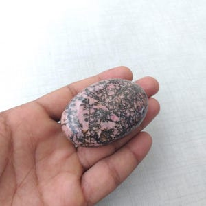 May include: A pink and black oval-shaped stone with a unique pattern of dark lines. The stone is smooth and polished, with a mix of pink and black colors. The stone is held in a hand.