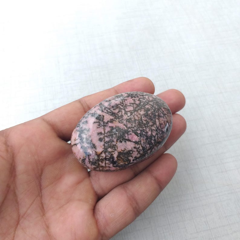 May include: A pink and black oval-shaped stone with a unique pattern of dark lines. The stone is smooth and polished, with a mix of pink and black colors. The stone is held in a hand.