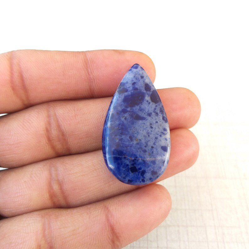 May include: A teardrop-shaped gemstone with a deep blue hue, speckled with lighter blue and dark spots. The polished surface reflects light, showcasing the natural patterns of the stone. The gemstone is held in a hand.