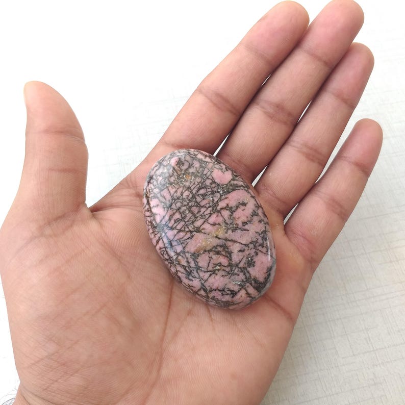 May include: An oval-shaped, polished gemstone with a pink base color and intricate black veining. The stone is held in an open hand, showcasing its smooth texture and natural patterns. The gemstone is a unique piece of natural decor.