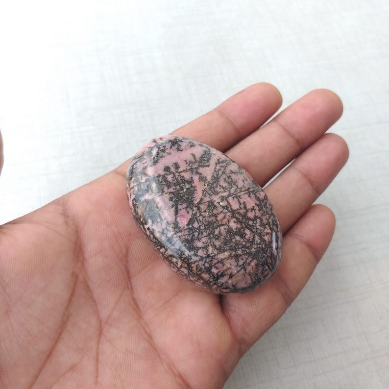 May include: An oval-shaped, polished stone with a pink base color and intricate black veining. The stone is held in a hand, showcasing its smooth texture and natural patterns. The stone's size is approximately 3 inches in length.