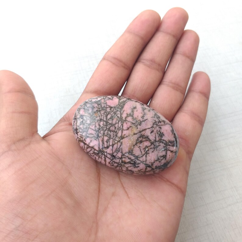 May include: A pink and black oval-shaped gemstone. The stone has a polished surface with a network of dark lines and veins. The gemstone is held in an open hand.