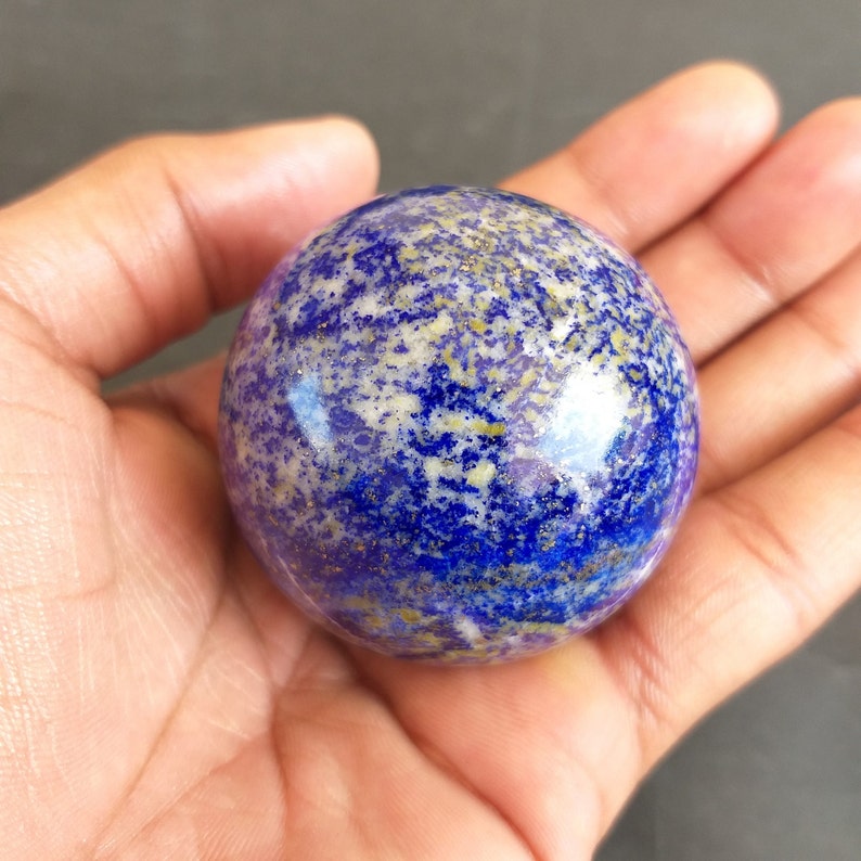 May include: A polished blue and gold lapis lazuli sphere, a type of gemstone, is held in a hand. The sphere has a speckled pattern of gold and white against a blue background.