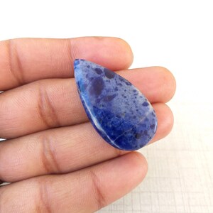 May include: A teardrop-shaped gemstone with a deep blue hue, speckled with lighter blue and dark spots. The polished surface reflects light, showcasing the natural patterns of the stone. The gemstone is held in a hand.