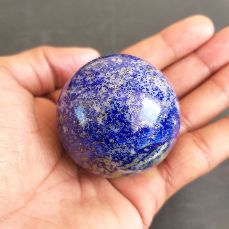 May include: A polished blue and gold lapis lazuli sphere, a type of gemstone, is held in a hand. The sphere has a smooth, shiny surface with a swirling pattern of blue and gold.