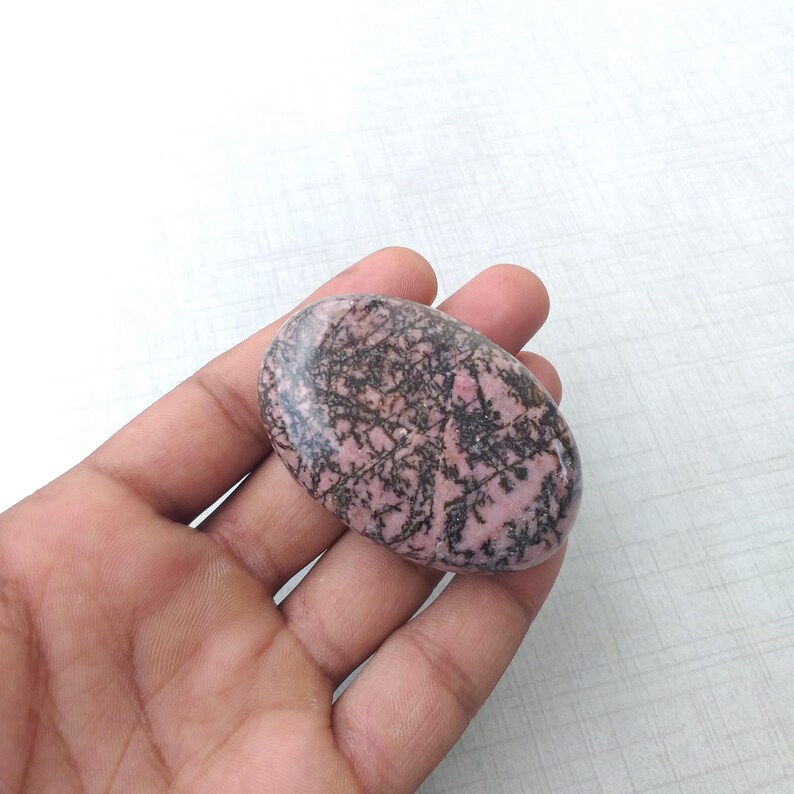 May include: Oval-shaped polished rhodonite stone. The stone is a soft pink color with intricate black veining patterns throughout. The stone is held in a hand against a white background. This natural stone is a unique piece for collectors or for use in jewelry.