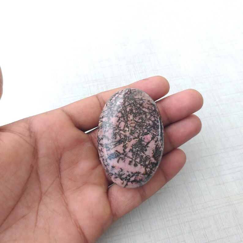 May include: An oval-shaped gemstone with a pink base and intricate black veining. The polished stone is held in a hand, showcasing its smooth surface and natural patterns. The gemstone's design is reminiscent of a delicate network.