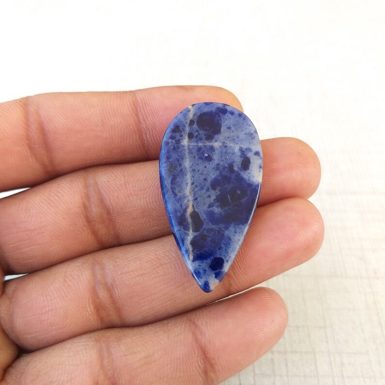 May include: A teardrop-shaped gemstone with a polished surface. The stone features a mix of deep blue and white hues, with darker blue spots and veins throughout. The gemstone is held in a hand, showcasing its size and shape.