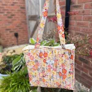 May include: A quilted tote bag featuring a vibrant floral pattern in shades of orange, yellow, and pink on a cream background. The bag has two matching fabric handles and a small white rectangular label with black text on the front centre.