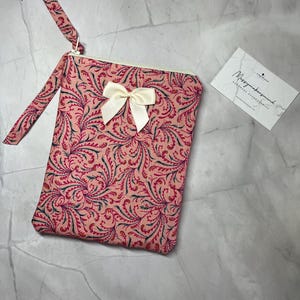 May include: A pink floral patterned clutch with a matching wrist strap and a cream-colored bow. The clutch has a zipper closure and a small tag that reads "Messymakersmind Handmade Accessories."