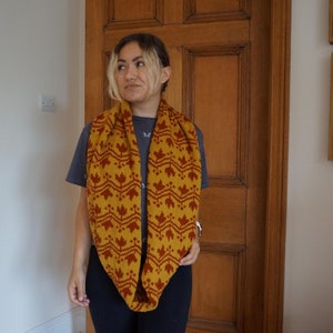 May include: A woman wearing a mustard yellow infinity scarf with a red leaf pattern. The scarf is made of a soft, knitted fabric.