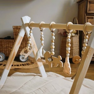 May include: A wooden baby gym with hanging toys. The frame is natural wood with white accents. The toys include wooden shapes and beads. A light-colored play mat is beneath the gym.