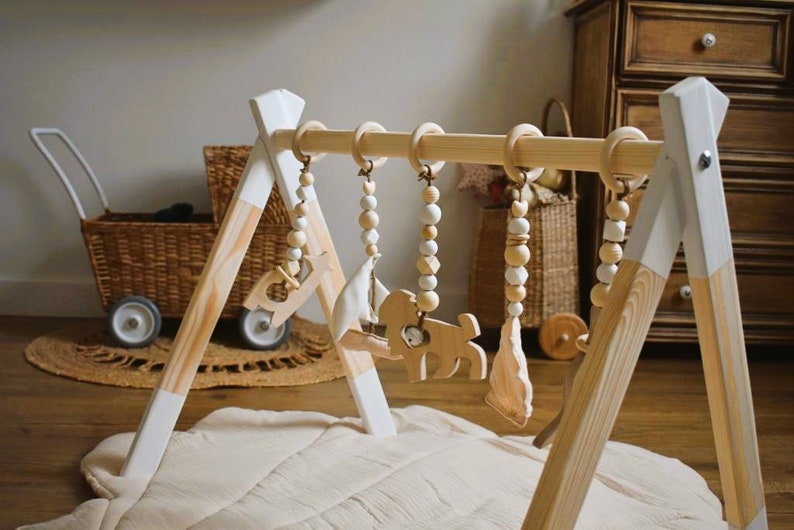 May include: A wooden baby gym with hanging toys. The frame is natural wood with white accents. The toys include wooden shapes and beads. A light-colored play mat is beneath the gym.