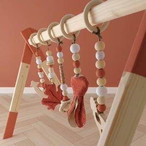 May include: A wooden baby gym with hanging toys. The frame is natural wood with red accents. Hanging toys include wooden beads, a leaf-shaped toy, and a red fabric toy with white polka dots. The gym is designed for infant play and development.