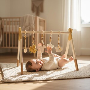 May include: Wooden baby play gym with hanging toys in a neutral nursery setting. The gym features a natural wood frame with yellow accents and a variety of wooden and felt toys. A baby is lying on a cream-colored rug, reaching for the toys.