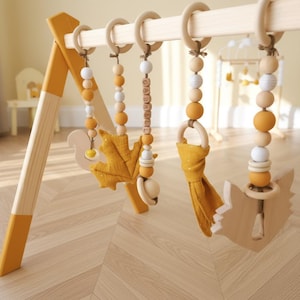 May include: Wooden baby play gym with hanging toys. The frame is natural wood with yellow accents. Hanging toys include wooden beads, a leaf, a squirrel, and a fox. The toys are in shades of yellow, white, and natural wood.