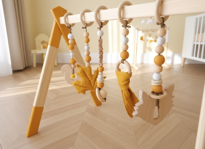 May include: Wooden baby play gym with hanging toys. The frame is natural wood with yellow accents. Hanging toys include wooden beads, a leaf, a squirrel, and a fox. The toys are in shades of yellow, white, and natural wood.