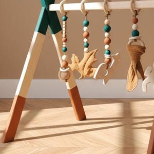 May include: Wooden baby play gym with hanging toys. The frame is painted in white, teal, and brown. The hanging toys include wooden beads, a felt leaf, a felt deer, and a fabric knot.
