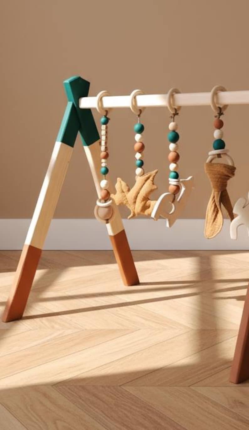 May include: Wooden baby play gym with hanging toys. The frame is painted in white, teal, and brown. The hanging toys include wooden beads, a felt leaf, a felt deer, and a fabric knot.