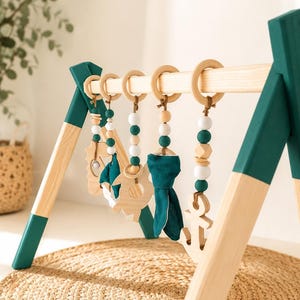 May include: Wooden baby play gym with hanging toys. The frame is natural wood with teal accents. Hanging toys include wooden shapes, teal fabric, and beads. The play gym is set on a woven mat.
