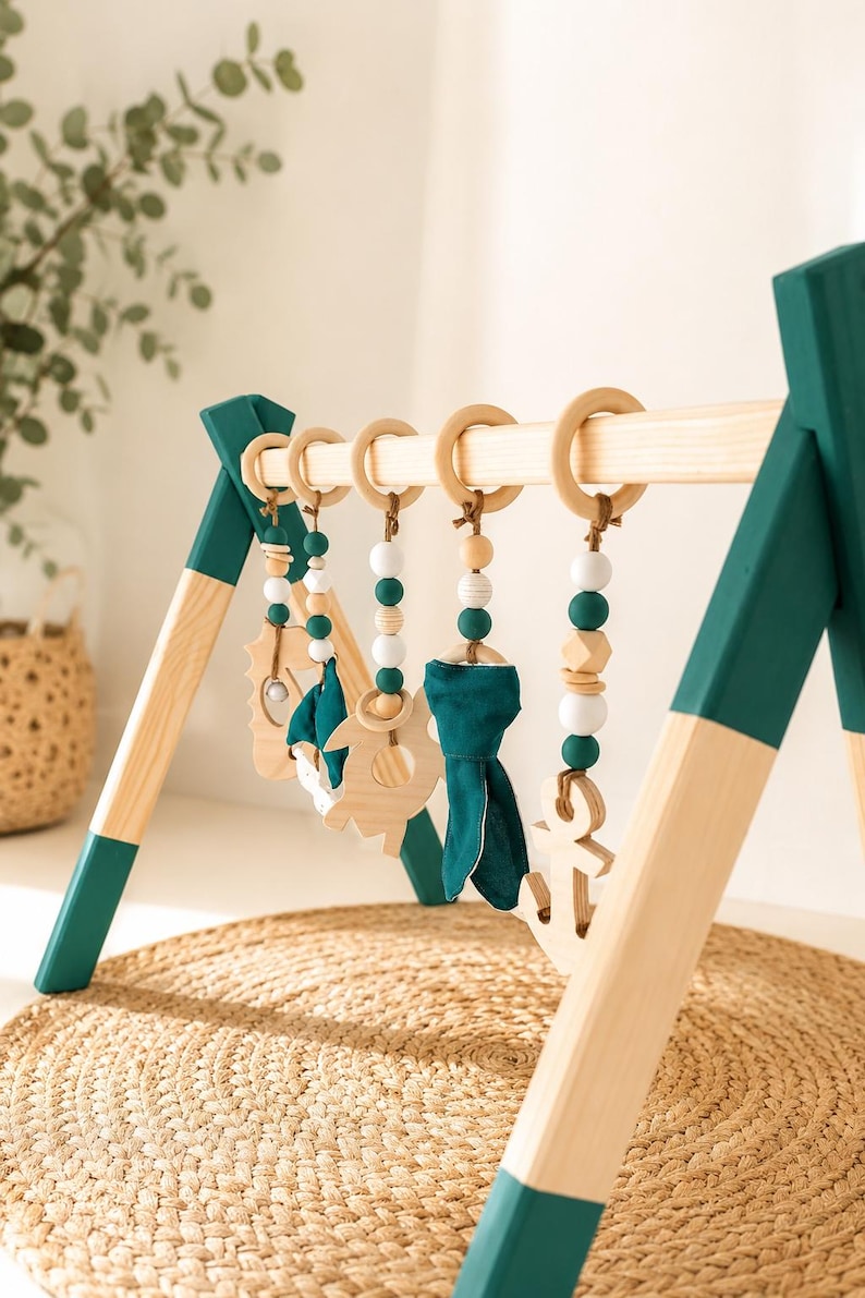 May include: Wooden baby play gym with hanging toys. The frame is natural wood with teal accents. Hanging toys include wooden shapes, teal fabric, and beads. The play gym is set on a woven mat.