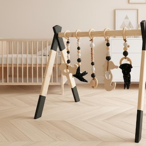 May include: A wooden baby play gym with hanging toys. The frame is natural wood with black accents. Hanging toys include wooden beads, a seahorse, a bird, and a seahorse-shaped teether. The background includes a crib and a dresser.