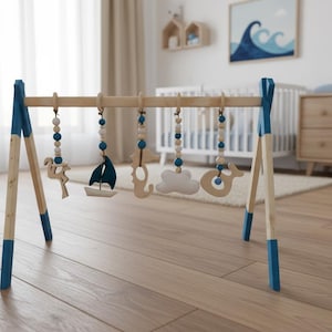 May include: Wooden baby play gym with blue accents and hanging toys. The toys include a sailboat, cloud, and whale. The gym is set up in a nursery with a crib and dresser in the background.