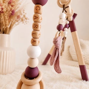 May include: Wooden baby gym with hanging toys. The toys include wooden beads, a ring, and a fabric element. The beads spell out the name "CARLA". The gym's legs are painted with a plum color.