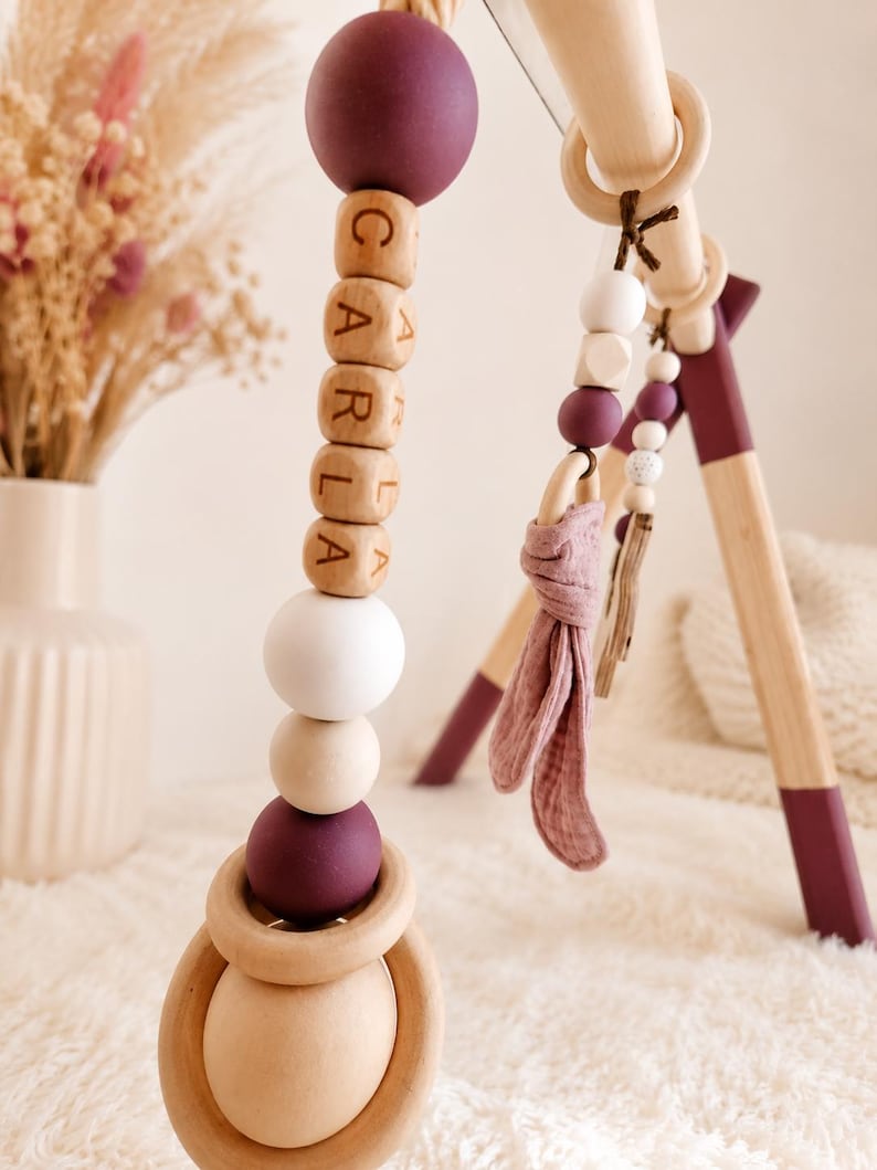 May include: Wooden baby gym with hanging toys. The toys include wooden beads, a ring, and a fabric element. The beads spell out the name "CARLA". The gym's legs are painted with a plum color.