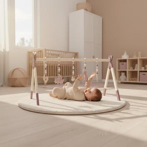 Könnte beinhalten: Ein Baby spielt unter einem Spielbogen aus Holz mit hängendem Spielzeug. Der Bogen hat einen hellen Holzrahmen mit lavendelfarbenen Akzenten. Das Baby liegt auf einer runden, cremefarbenen Spielmatte. Ein Kinderbett und andere Möbel sind im Hintergrund zu sehen.