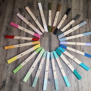 May include: A collection of 23 wooden planks, each painted with a different color and numbered. The planks are arranged in a circular pattern on a wooden surface. The colors include white, pink, red, yellow, green, blue, and gray.