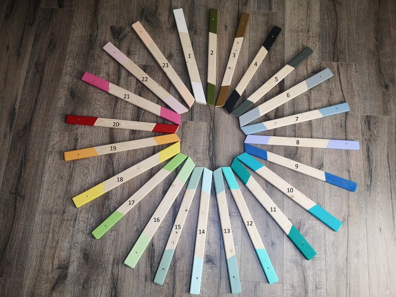 May include: A collection of 23 wooden planks, each painted with a different color and numbered. The planks are arranged in a circular pattern on a wooden surface. The colors include white, pink, red, yellow, green, blue, and gray.