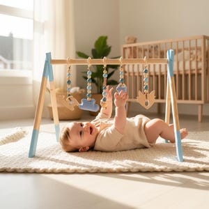 May include: A wooden baby play gym with blue accents and hanging toys. The toys include wooden shapes and beaded strands in shades of blue and natural wood. A baby is lying on a cream-colored rug, reaching for the toys.