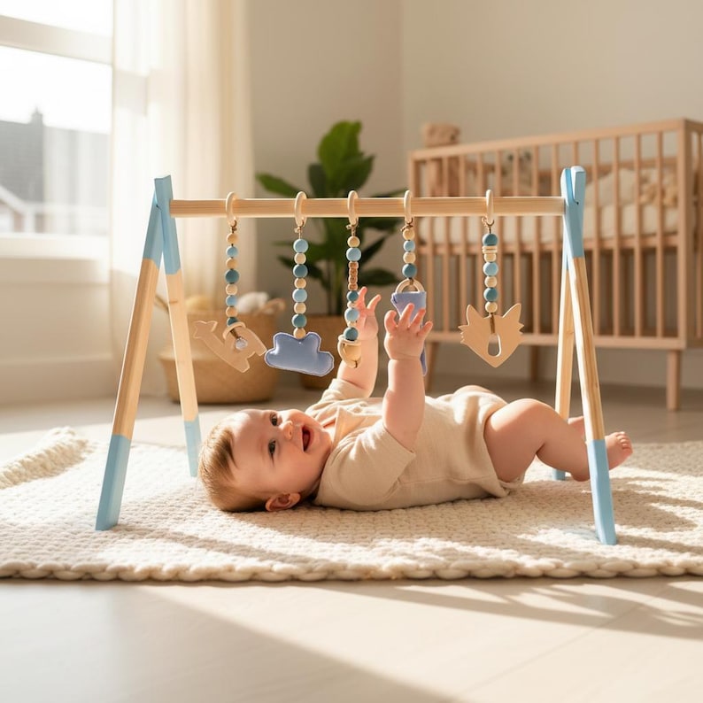 May include: A wooden baby play gym with blue accents and hanging toys. The toys include wooden shapes and beaded strands in shades of blue and natural wood. A baby is lying on a cream-colored rug, reaching for the toys.