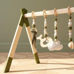 May include: A wooden baby play gym with a natural wood finish and olive green accents. Hanging from the wooden bar are wooden beads, a whale-shaped toy, a cloud-shaped toy, and a wooden ring with a fabric accent.