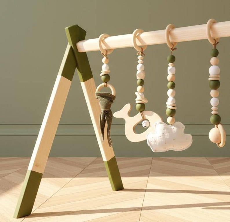 May include: A wooden baby play gym with a natural wood finish and olive green accents. Hanging from the wooden bar are wooden beads, a whale-shaped toy, a cloud-shaped toy, and a wooden ring with a fabric accent.