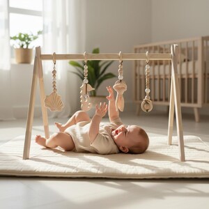 May include: A wooden baby play gym with hanging toys, including a seashell, seahorse, and other shapes. A baby is lying on a cream-colored mat, reaching for the toys. The gym is made of light-colored wood.