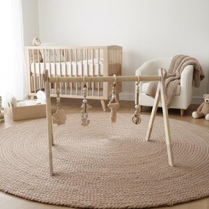 May include: A wooden baby gym with hanging toys is set up on a round, woven jute rug. The nursery scene includes a crib, a white dresser, a teddy bear, and a potted plant. The room has a neutral color palette.