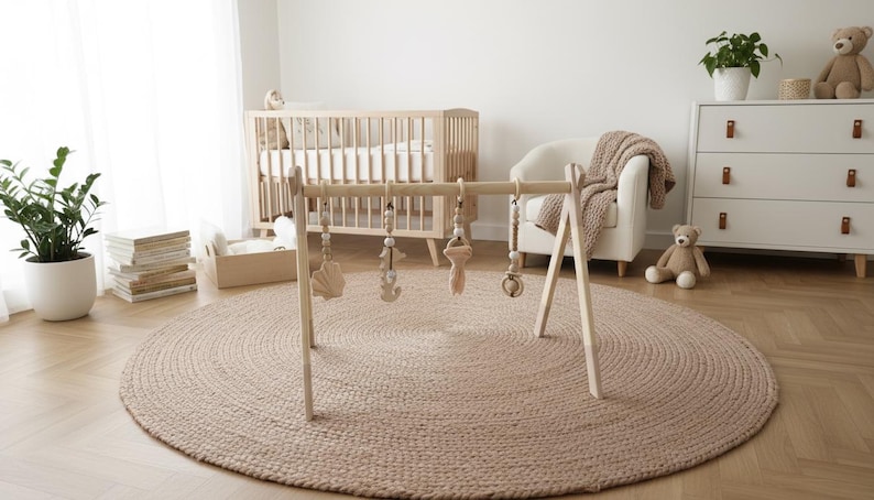 May include: A wooden baby gym with hanging toys is set up on a round, woven jute rug. The nursery scene includes a crib, a white dresser, a teddy bear, and a potted plant. The room has a neutral color palette.