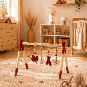 Peut inclure: Un portique d'éveil en bois composé d'une structure en bois naturel avec des pieds aux extrémités peintes en rouge foncé. Cinq jouets suspendus incluent des perles en bois, une feuille en tissu rouge, un renard en tissu et des formes en bois.