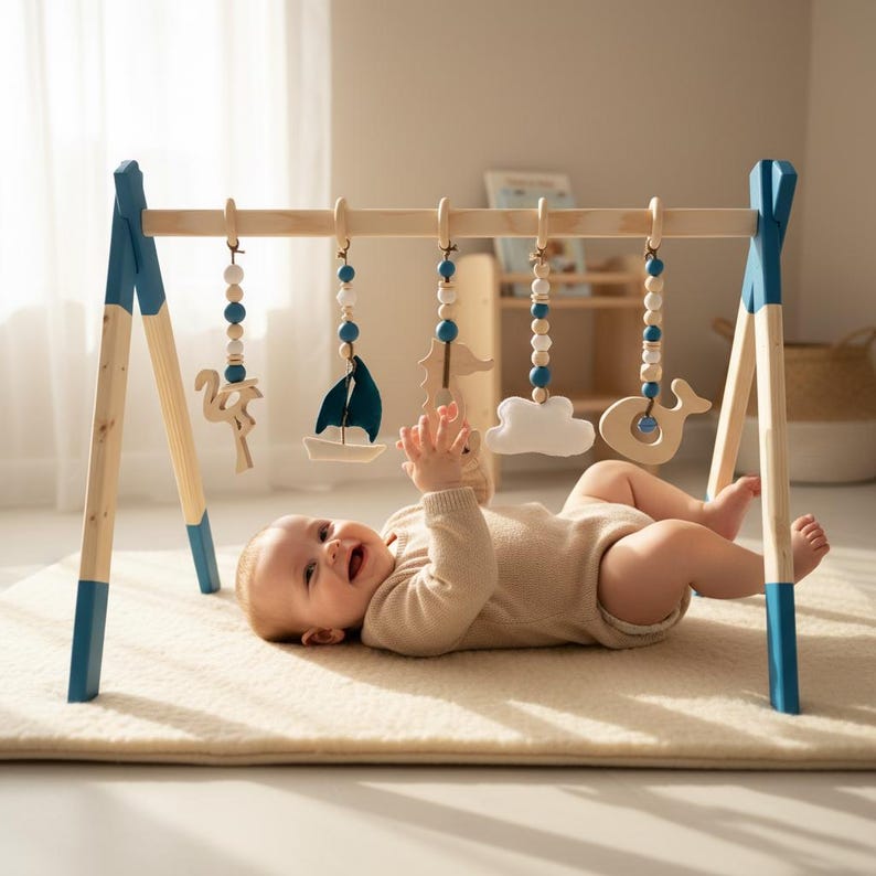 May include: A wooden baby play gym with blue accents and hanging toys. The toys include a sailboat, cloud, whale, and wooden beads. A baby in a beige onesie is lying on a cream-colored rug, reaching for the toys.
