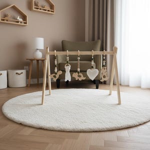 May include: A wooden baby gym with hanging toys, including a heart, dinosaur, and bear, sits on a white, round rug. The room features a crib, shelves, and a chair, all in neutral tones, creating a calming nursery environment.
