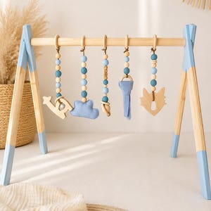 May include: Wooden baby play gym with blue accents and hanging toys. The toys include a wooden airplane, a blue cloud, a wooden bear, a blue key, and a wooden fox. The wooden frame has blue painted legs and a natural wood top.
