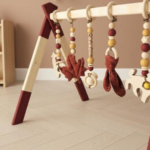 May include: Wooden baby play gym with hanging toys. The frame is natural wood with burgundy accents. Hanging toys include wooden beads, a leaf, a ring with a fabric tie, and a wooden animal. The background includes a wooden shelf with toys.