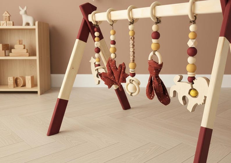 May include: Wooden baby play gym with hanging toys. The frame is natural wood with burgundy accents. Hanging toys include wooden beads, a leaf, a ring with a fabric tie, and a wooden animal. The background includes a wooden shelf with toys.