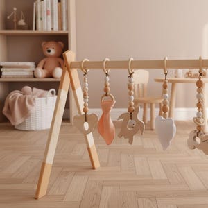 May include: Wooden baby play gym with hanging toys. The toys include a heart, a cloud, a fish, and a turtle. The gym is made of light-colored wood with a natural finish. The toys are in neutral colors, with some peach and white accents.