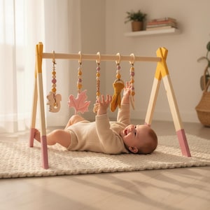 May include: A wooden baby play gym with hanging toys. The frame is painted with yellow and pink accents. The toys include a wooden squirrel, a pink leaf, and other colorful shapes. A baby is lying on a cream-colored rug, reaching for the toys.