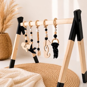 May include: A wooden baby play gym with hanging toys. The frame is natural wood with black accents. The hanging toys include wooden shapes, black and white beads, and wooden rings. The play gym is set on a woven mat.